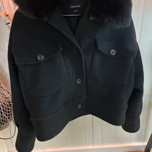 Svart jacka från Meotine - Snygg svart jacka från Meotine med stor pälskrage, två stora bröstfickor med knappar och markerad midja. Jackan är i ullblandning och har en boxig, lite oversized passform. 