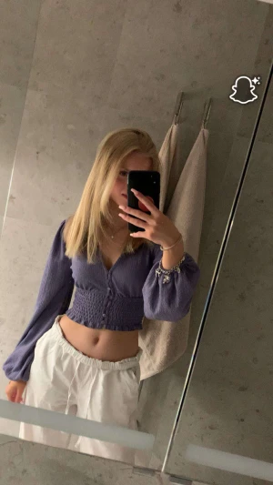 Lila croppad blus med knappar - Säljer en lila blus med croppad passform och långa puffärmar. Toppen har v-ringning och knappar framtill samt smockad midja som ger en snygg siluett. Perfekt för dig som gillar trendiga och bekväma plagg med lite extra detaljer.