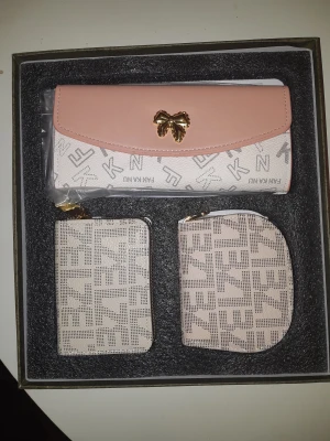 Set med plånbok och småväskor FANANAU - Snyggt set med en plånbok och två mindre väskor från FANANAU. Plånboken har ljusrosa lock med guldfärgad rosett och mönstrad vit botten. De små väskorna är beige med grafiskt mönster och dragkedja. Perfekt för att hålla koll på kort och småsaker.