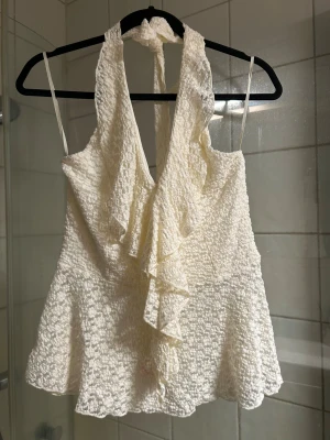 Beige volangblus från Missäy - Supersöt beige blus från Missäy med volangdetaljer framtill och halterneck. Blusen är ärmlös och har ett spetsliknande, blommigt tyg som ger en romantisk vibe. Perfekt att styla med jeans eller kjol för en trendig look.