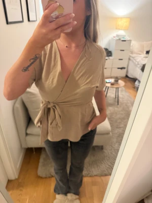 Beige omlottopp med knyt i midjan - Säljer en beige omlottopp med korta ärmar och v-ringning. Toppen har ett snyggt knyt i midjan som ger en feminin siluett. Materialet känns mjukt och lätt, perfekt för vår och sommar. Passformen är normal och toppen är enkel att matcha med jeans eller kjol.