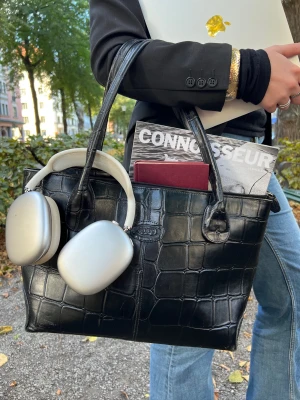 Svart croco handväska - Snygg svart handväska i croco-mönstrat skinn. Väskan har handtag och rymligt fack som rymmer laptop, magasin och hörlurar. Perfekt för dig som vill ha en stilren och lyxig look med lite edge.