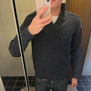 Mörkblå stickad tröja Industrialize - Mörkblå stickad tröja från Industrialize med hög krage och tre stora knappar vid halsen. Tröjan har lång ärm, ribbade muddar och en klassisk, lite loose passform. Perfekt för dig som gillar stilrena och bekväma plagg.