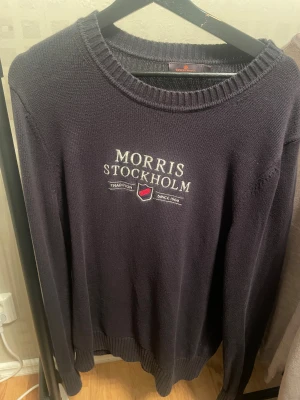 Mörkblå stickad tröja Morris M - Snygg mörkblå stickad tröja från Morris Stockholm i storlek M. Tröjan har broderad logga och text på bröstet, rund hals och ribbade muddar vid ärmslut och nederkant. Perfekt för dig som gillar klassisk stil med en modern touch.
