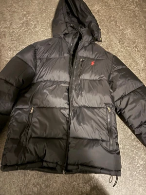 Svart pufferjacka från Ralph Lauren - Svart pufferjacka från Ralph Lauren med huva och dragkedja. Jackan har två fickor med dragkedja och en röd broderad logga på bröstet. Perfekt för kalla dagar och riktigt snygg streetstil.använd en gång i typ 20 min men annars ny