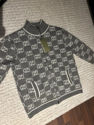 Grå stickad kofta från Gucci - Säljer en grå stickad kofta från Gucci med hel dragkedja och GG-mönster över hela tröjan. Koftan har vita detaljer vid muddar och krage samt två fickor framtill. Perfekt för dig som vill ha en snygg och lyxig look.