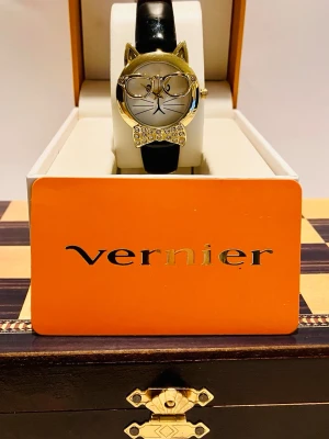 Svart och guld kattklocka från Vernier - Unik klocka med kattmotiv från Vernier, har guldfärgat boett med öron och glasögon, samt en glittrig rosett med strass. Svart läderband och detaljerad urtavla med kattansikte. Perfekt för dig som gillar coola accessoarer med personlig touch.