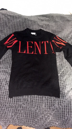 Svart stickad tröja från Valentino - Svart stickad tröja från Valentino med röd logotyp och grafiska detaljer på ärmarna. Rund halsringning och ribbade muddar vid ärmslut och nederkant. Snygg och stilren design som sticker ut med de röda accenterna.