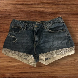 Blå jeansshorts med spets från 157 - Snygga blå jeansshorts från 157 i M med coola slitningar och vit spetskant längst ner. Perfekt för dig som gillar att sticka ut med detaljer och vill ha en unik look på sommaren. ALDRIG ANVÄNDA 