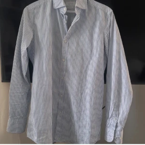 Randig skjorta från Massimo Dutti - Snygg skjorta från Massimo Dutti med smala blå och vita ränder. Klassisk krage, knappar framtill och långa ärmar med manschett. Skjortan är i mjuk bomull och har en normal passform som sitter schysst på kroppen.