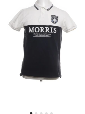 Svartvit piké från Morris, storlek M - Säljer en snygg pikétröja från Morris i storlek M. Toppen är svart och vit med kontrastfärgade partier, korta ärmar och klassisk krage med ränder. Framsidan har Morris-logga och tryck. Materialet är mjuk bomull, perfekt för en chill och sportig look.
