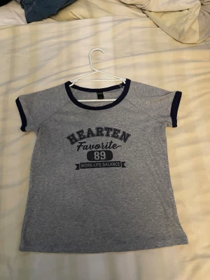 Grå t-shirt med blå detaljer från Dazy - Snygg grå t-shirt från Dazy med mörkblå kant vid hals och ärmslut. Tryck på bröstet med texten 'HEARTEN Favorite 89 WORK-LIFE BALANCE'. T-shirten har korta ärmar och en klassisk rund hals. Perfekt för en chill och sportig look.