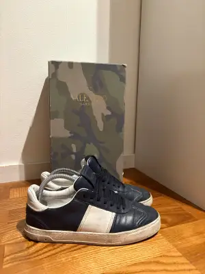 Snygga Valentino Garavani sneakers i mörkblått och vitt skinn med rund tå och platt sula. Klassisk design med svarta snören och coola nitar på sulan. Kommer med originalkartong i camouflage-mönster. Perfekt för dig som gillar exklusiva streetwear-vibbar.