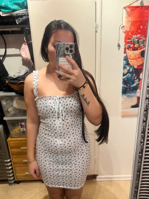 Klänning SHEIN  - Säljer en söt vit kortklänning med blått blommigt mönster och snörning framtill. Klänningen har volangdetaljer på axelbanden och en figurnära passform. Perfekt för sommarens alla tillfällen, superfin och bekväm!