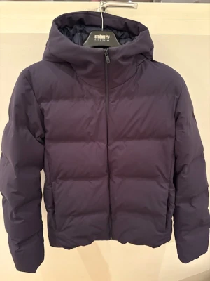 Mörkblå pufferjacka från Uniqlo - Säljer en stilren mörkblå pufferjacka från Uniqlo med huva och dragkedja framtill. Jackan är vadderad och har en clean look utan synliga detaljer eller mönster. Perfekt för kalla dagar och enkel att matcha med din vardagsstil.