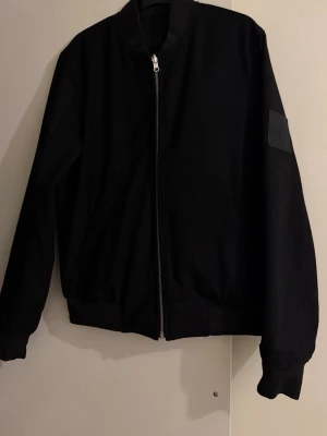 Svart bomberjacka från Peak Performance - Svart bomberjacka från Peak Performance med stilren design, ribbade muddar och silverfärgad dragkedja. Jackan har två stora fickor framtill och en diskret logopatch på ärmen. Materialet är slitstark polyester och passformen är normal. Jackan är väldigt unik och är in & ut vändig vilket ger dig två jackor i en jacka!