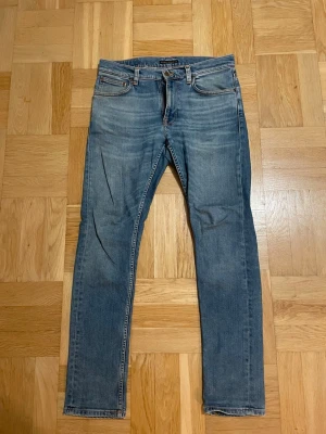 Blå jeans från Nudie Jeans, 30/32 - Snygga blå jeans från Nudie Jeans med klassisk femficksdesign och orangea sömmar. Modellen har raka ben och normal passform, med cool broderad detalj på bakfickorna. Perfekt för dig som gillar stilrena och tidlösa jeans. Jeansen har ett hål i skrevet men som går att sy igen. Hör av dig vid frågor. Mvh Ville