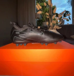 Nike svarta fotbollsskor med dobbar - Säljer ett par svarta Nike fotbollsskor med snygga silverdetaljer och snörning. Skorna har rund tå och platt sula med dobbar för bra grepp på planen. Tillverkade i syntetmaterial som är slitstarkt och lätt att rengöra. Perfekt för dig som vill ha stilrena och funktionella skor till träningen.