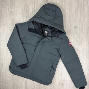 Canada Goose macmillan äkta - Snygg mörkgrå dunjacka från Canada Goose med huva och klassisk logga på ärmen. Jackan har dragkedja och knappar framtill, samt två sidofickor. Perfekt för kalla dagar och har en stilren, sportig look. Har äkthetsbevis