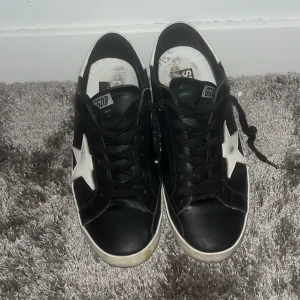 Svarta sneakers med vit stjärna GGDB - Svarta sneakers från Golden Goose med vit stjärndetalj på sidan och vit sula. Skorna har snörning och rund tå, tillverkade i skinn med klassisk låg profil. Perfekta för dig som gillar streetstyle och vill sticka ut med en ikonisk design.