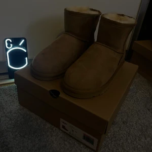 UGG Classic Mini II boots beige - Sprillans nya Uggs! UGG Classic Mini II boots i beige mocka med rund tå och platt sula. Fodrade med mjuk ull för extra värme och komfort. Ikonisk UGG-logga bak på hälen och klassisk siluett som är perfekt för kyliga dagar.