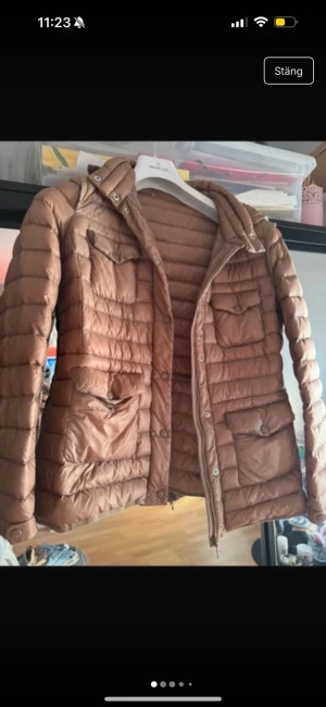 Brun Moncler dunjacka, storlek 2 - Säljer en brun dunjacka från Moncler med klassisk quiltad design, flera fickor framtill och logotyp på ärmen. Jackan har lång ärm, dragkedja och knappar framtill. Perfekt för kyliga dagar och riktigt snygg till streetwear eller casual outfits.