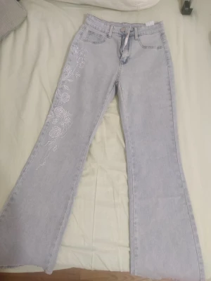 Ljusblå jeans med brodering - Snygga ljusblå jeans med vita blombroderier längs ena benet. Modellen har hög midja och vida ben. Helt nya, aldrig använt. Kan passa även någon som bär 34.