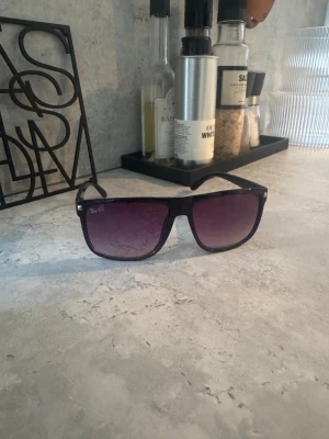 Svart Ray-Ban solglasögon - Snygga svarta Ray-Ban solglasögon med breda bågar och rektangulär form. Glasen har en mörk toning som går från lila till svart, vilket ger en cool vibe. Perfekt accessoar för att lyfta din stil under soliga dagar.