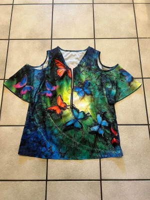 Färgglad topp med fjärilar och dragkedja - Säljer en unik topp med färgstarkt fjärilsmönster i blått, grönt, rött och rosa. Toppen har korta ärmar med öppna axlar och en snygg ringformad dragkedja framtill. Materialet är mjukt och syntetiskt, perfekt för dig som gillar att sticka ut med coola prints.
