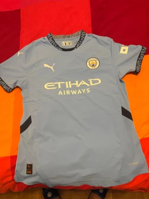 Manchester City Puma Authentic fotbollströja L - Manchester Citys officiella matchtröja från Puma i ljusblått med klubbmärke på bröstet och Etihad Airways-tryck. Tröjan har svarta detaljer på sidorna och mönstrad krage. Tillverkad i ventilerande syntetmaterial för maximal komfort på planen.