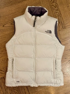 Vit dunväst från The North Face XS - Snygg vit dunväst från The North Face med hög krage och grått foder. Västen har dragkedja framtill, två sidofickor med dragkedjor, innerficka med dragkedja och broderad logga på bröstet och ryggen. Perfekt för lager-på-lager och chill stil. 