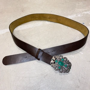 Brunt skärp sillverspänne Humana - Brunt bälte i skinn med ett stort, dekorativt silverfärgat spänne prytt med gröna stenar. Defekt: lossnat lite i kanten på skinnet- går att limma. Mått: 118 cm. 