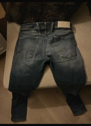 Replay blå jeans med raka ben - Snygga blå jeans från Replay med klassisk femficksdesign och raka ben. Jeansen har ljusa slitningar framtill och bak, samt tydliga sömmar på bakfickorna. Märkespatch i beige på linningen och metallknapp framtill. Perfekt för dig som gillar en stilren och tidlös look.