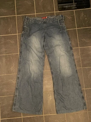 Blå wide jeans med snörning i sidan  - Säljer ett par blå jeans med breda ben och cool snörning längs båda sidorna vid midjan. Jeansen har en avslappnad passform och är tillverkade i klassiskt denimtyg. Perfekt för dig som gillar en lite retro och unik stil.