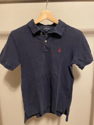 Mörkblå pikétröja från Polo Ralph Lauren - Snygg mörkblå pikétröja från Polo Ralph Lauren med klassisk krage och två knappar framtill. Tröjan har korta ärmar och den ikoniska röda broderade loggan på bröstet. Materialet är mjuk bomull som känns skönt mot huden.