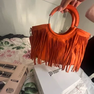 Orange handväska med fransar - Cool orange handväska med runda metallhandtag och långa fransar på framsidan. Väskan har ett färgglatt mönstrat innerfoder och en avtagbar axelrem. Perfekt för dig som vill sticka ut med en unik accessoar.