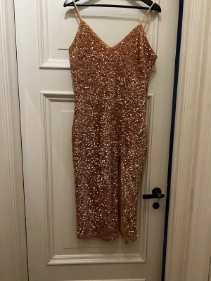Glittrig klänning i guld - Säljer en snygg klänning i stretch med smala axelband, täckt av glittriga paljetter i guld. Modellen har v-ringad hals. Materialet är stretch syntet och paljett, med en figurnära passform. 