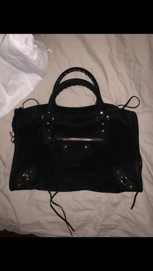 Svart Balenciaga väska i mocka - Säljer en svart handväska från Balenciaga i lyxig mocka med snygga detaljer i guld och dragkedjor. Väskan har både handtag och axelrem, samt flera fack och innerfickor. Klassisk design med flätade handtag och  metalldetaljer. Väskan är i mycket gott skick, med undantag för en mindre skada på ett av handtagen där mockan är något nött. Kvitto, dustbag och tags tillkommer.