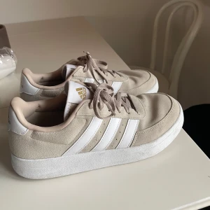 Beige Adidas sneakers med mockadetaljer - Snygga beige sneakers från Adidas med klassiska vita ränder på sidorna och guldfärgad logga på plösen och hälen. Ovandelen är i mocka och syntet, med vita snören och platt sula. Perfekta för en clean och stilren look.