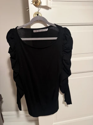 Svart blus med volangärmar Tiger of Sweden - Svart blus från Tiger of Sweden i storlek S. Modellen har långa ärmar med snygga volangdetaljer längs axlarna och ärmarna. Tunn och mjuk kvalitet med rund halsringning 🖤