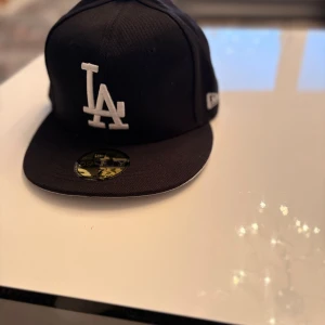 Svart LA-keps New Era 59FIFTY - Säljer en helt ny svart New Era 59FIFTY keps med LA-logga i vitt framtill. Klassisk flat brim och broderad logga på sidan. Har en stilren look som passar perfekt till streetwear. Märkesetikett och originalstickers finns kvar. 