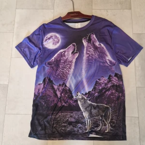 Lila t-shirt med vargar och måne - Säljer en unik t-shirt med ett stort tryck av tre ylande vargar under en fullmåne och berg i bakgrunden. T-shirten är lila med inslag av svart och har en cool naturinspirerad design. Modellen är kortärmad och materialet känns som polyester, perfekt för dig som gillar statement-plagg.