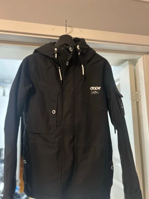 Svart vindjacka från Dope Snow S - Svart vindjacka från Dope Snow med huva och vita snören. Jackan har flera fickor med tryckknappar, logga på bröstet och ärm, samt coola detaljer som metallöljetter. Perfekt för dig som vill ha en varm å skön jacka nu i vinter till skid/snowbourd säsongen. Snygg och praktisk design. Jag har använt den 1 säsong. Jackan är i storlek S. Jag är 172cm lång som har haft den. Nypris 2299kr 
