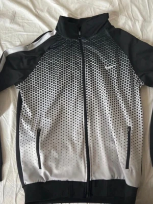 Svart och vit Nike Dri-Fit jacka - Nike Dri-Fit jacka med svart och vit färg, coolt prickigt mönster framtill och helsvart baksida. Jackan har dragkedja, ståkrage och två fickor med dragkedja. Perfekt för träning eller chill. Snygg logga på bröstet och ryggen.