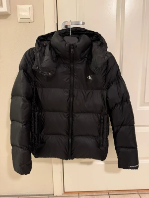 Svart pufferjacka från Calvin Klein - Svart pufferjacka från Calvin Klein med huva och dragkedja framtill. Jackan har CK-logga på bröstet och Calvin Klein-brodyr på ärmen. Använd 1 säsong så i mycket gott skick. Nypris 2500kr. 