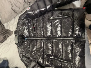 Sprillans ny Svart Moncler pufferjacka herr - Säljer en svart pufferjacka från Moncler med glansig finish och klassisk logga på ärmen. Jackan har flera dragkedjeförsedda fickor och quiltad design. Perfekt för kalla dagar och riktigt snygg till streetwear-stil.