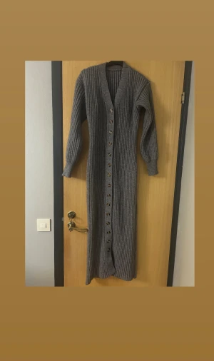Grå lång stickad klänning med knappar - Säljer en lång, ribbad stickad klänning i grå färg med v-ringning och knappar hela vägen framtill. Klänningen har långa ärmar och är stickad i ett mjukt material som ger en chill och cozy vibe. Den går även att knäppa upp och ha som kofta. En väldigt stretchig klänning som funkar i större strlk än bara M