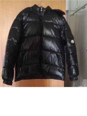 Svart Moncler dunjacka med huva - Säljer en svart, glansig dunjacka från Moncler med huva och dragkedja framtill. Jackan har klassisk pufferstil, Moncler-logga på ärmen och praktiska fickor med dragkedja. Perfekt för kalla dagar när du vill hålla stilen och värmen.