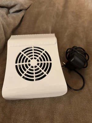 Vit nageldammsugare - Elektrisk nageldammsugare i vit plast med rund fläkt och galler, perfekt för att suga upp damm vid nagelfilning. Kommer med strömadapter och har en enkel on/off-knapp på sidan. Smidig storlek som passar på nagelbordet. 