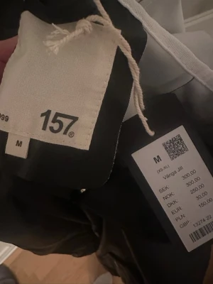 Svart regnjacka med huva från 157 - Svart regnjacka från 157, modell VÅNGA JILL. Jackan har huva med vit snörning, tryckknappar framtill och två fickor med knappstängning. Materialet är syntet och insidan är ljus. Perfekt när det regnar och du vill hålla dig torr.